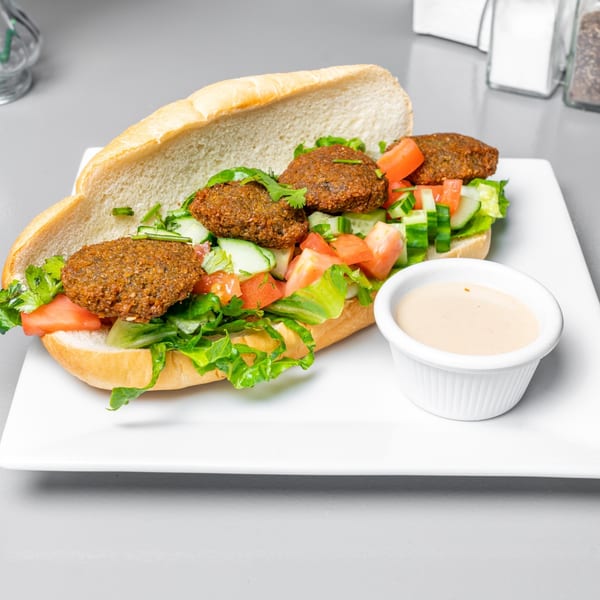 A King of Koshary falafel sandwich with lettuce, tomatoes, cucumbers, and a side of sauce.